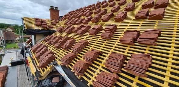 Vue large d'une toiture en cours de réfection, tuiles neuves réparties par piles sur le liteaunage jaune