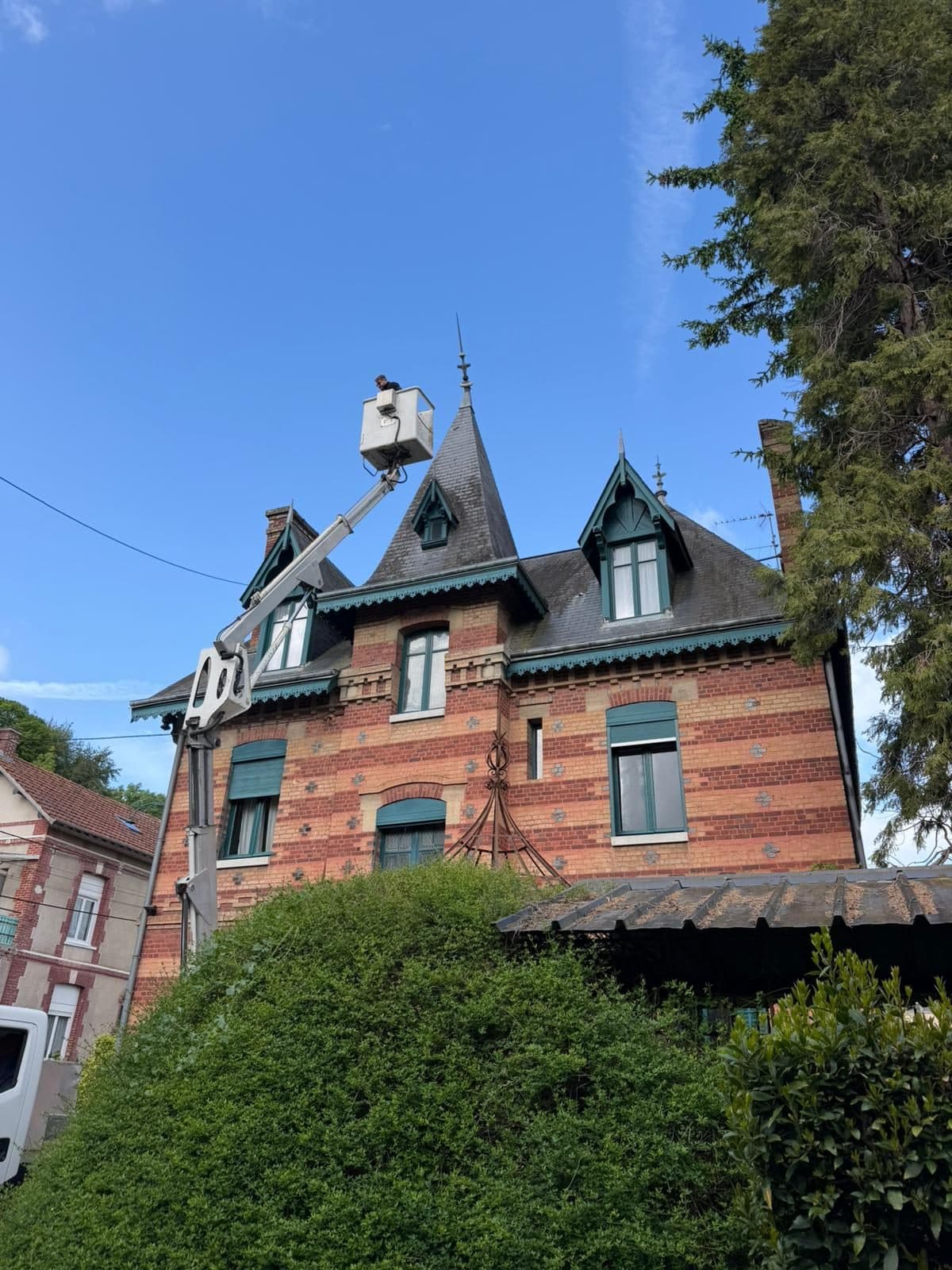 Couvreur en nacelle sur la toiture en ardoise d'une maison de maître en brique, avec tourelle, lucarnes ouvragées et épi de faîtage