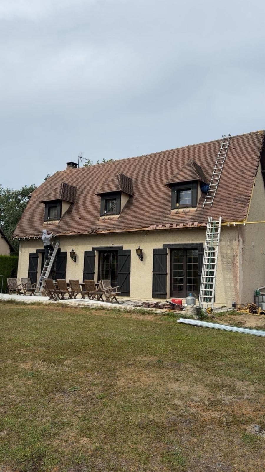 Rénovation de la couverture en tuiles d'un pavillon à trois lucarnes, couvreur sur échelle au niveau de l'égout et tuiles neuves stockées au sol
