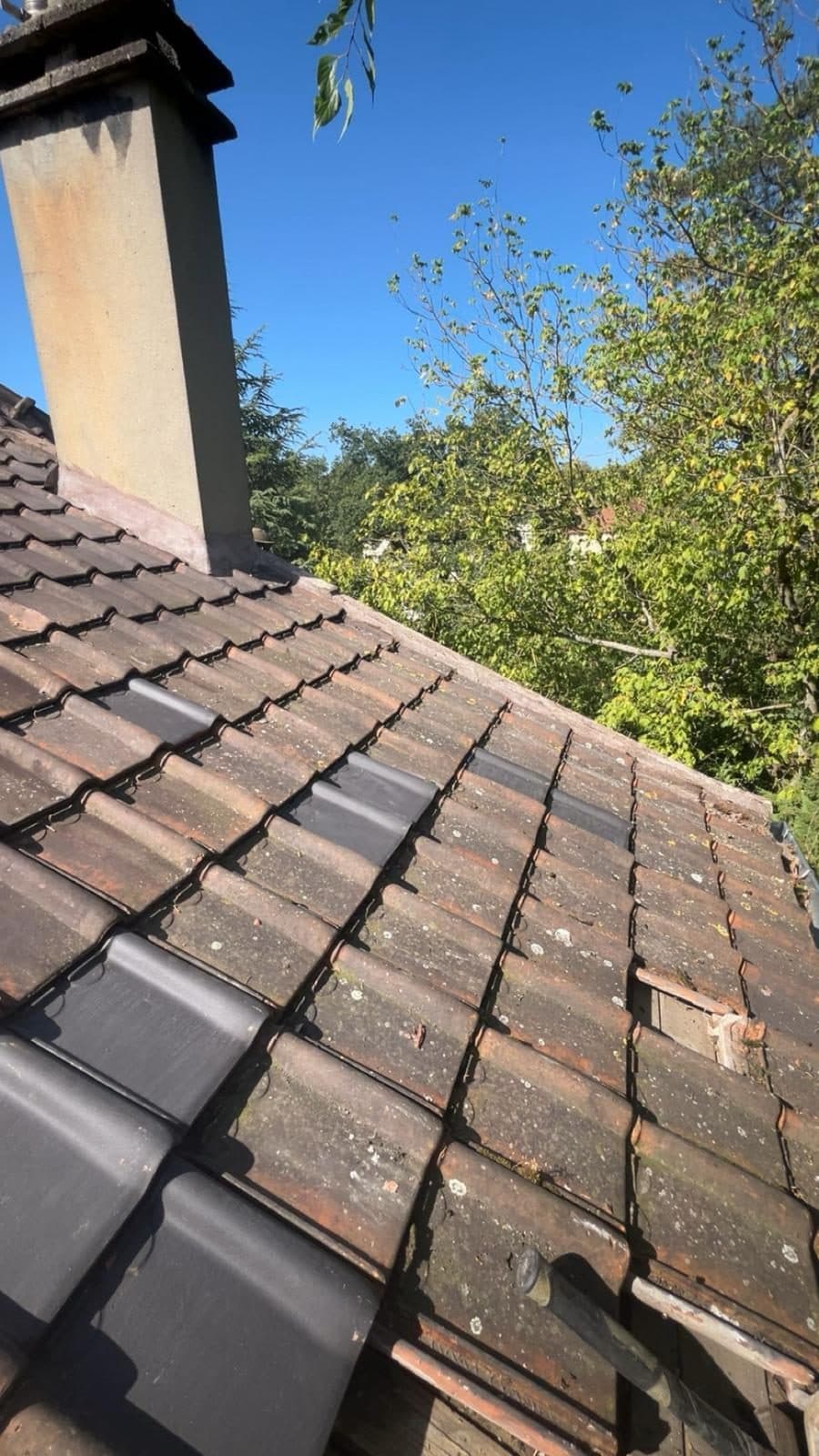 Tuiles neuves posées en remplacement d'éléments cassés sur une toiture ancienne couverte de lichen, près d'une souche de cheminée